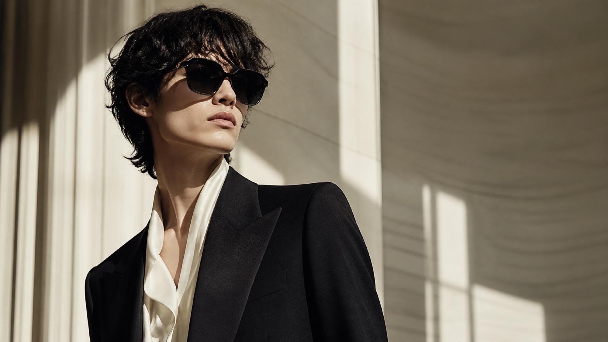 A person with tousled dark hair in a sharp black tuxedo and cream silk neck-tie blouse, photographed in dramatic side light, wearing oversized geometric sunglasses with a subtle hexagonal cut.