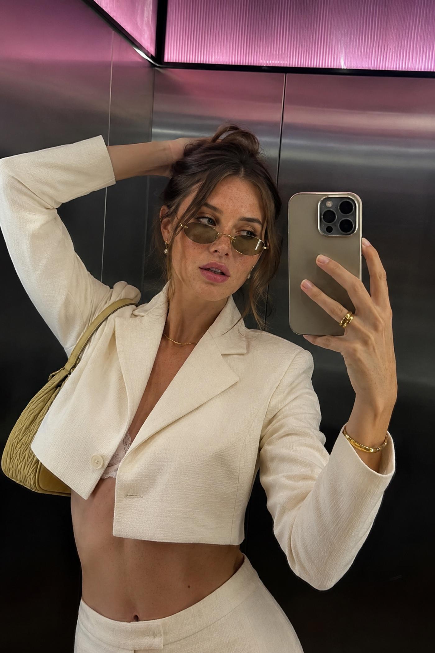 Woman in slim gold-frame oval sunglasses with green-tint lenses, taking an elevator mirror selfie.
