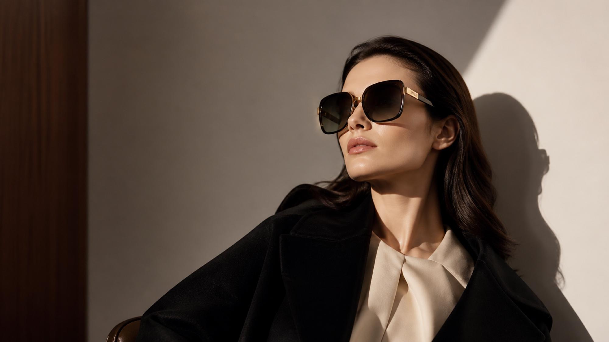 A woman in a black coat and cream blouse wearing oversized squared gold-edged sunglasses, photographed against a wall in dramatic side light.