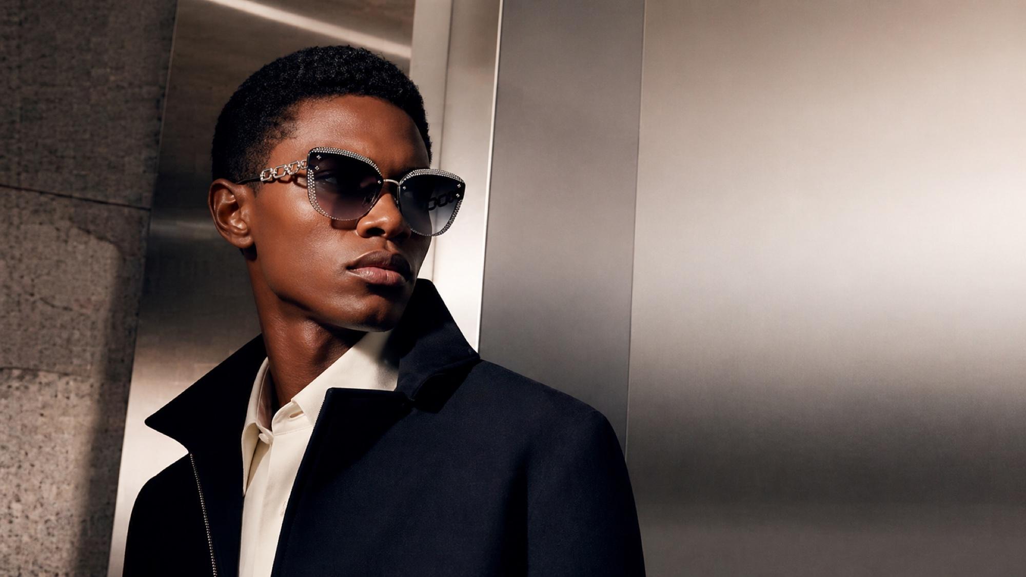 A man with deep skin in a navy coat and cream shirt, photographed against a metallic interior, wearing ornate butterfly-shaped sunglasses with rhinestone and silver detail.