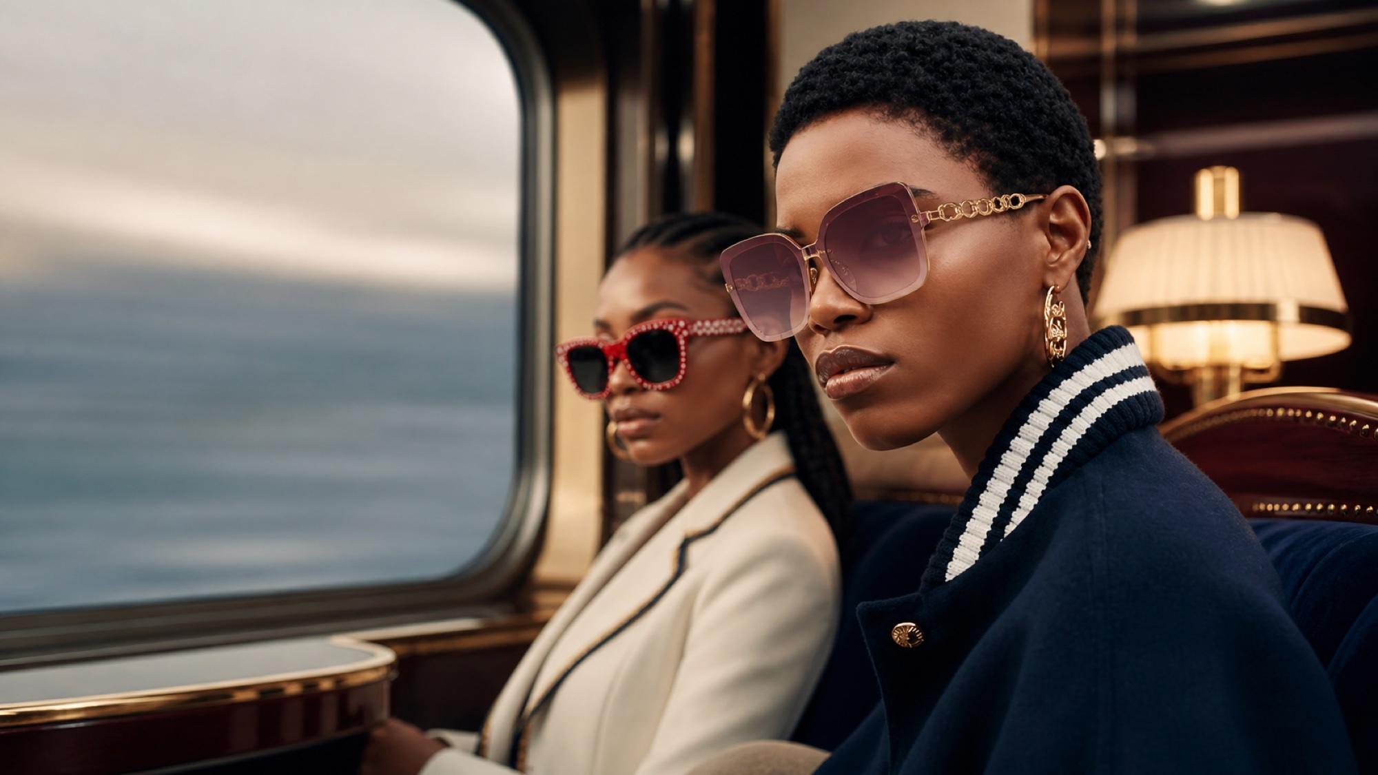 Two women on a luxury train, one wearing rose-pink oversized sunglasses with a gold chain detail on the temples, the other wearing red rhinestone-studded frames.